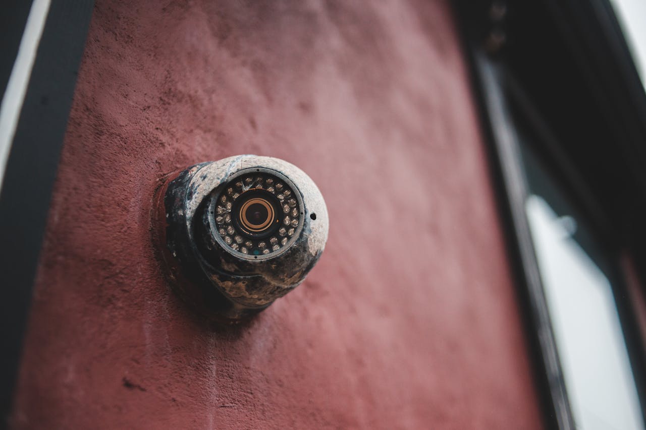 Close-up of an old security camera mounted on a weathered red wall, capturing a rustic exterior view.